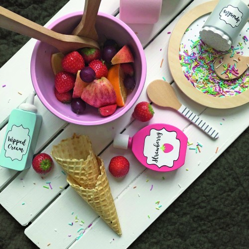 Serviervorschlag für das Fruchtsalat-Set JB-W7161: Rosa Schüssel mit Spielzeug-Früchten, Eiswaffeln und Saucenflaschen auf einem weißen Holztisch